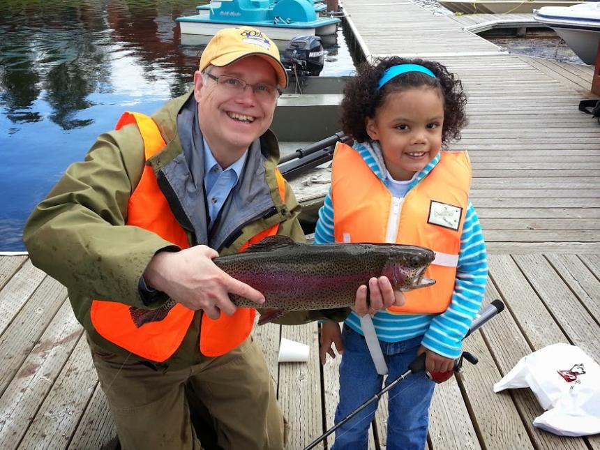 Volunteer holds a fish that was caught at youth fishing event, with young child standing nearby. 