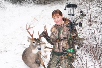 Hunter holding a bow kneeling next to a harvested deer in the snow