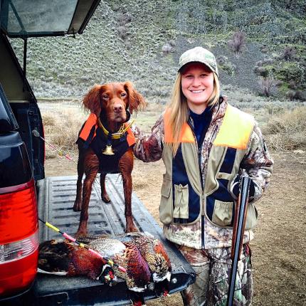 Photo of a hunter holding her shotgun and petting her dog with three harvested pheasants.