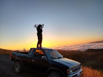 Photo of a bow hunter standing on the roof of a pickup truck lookout out over a scenic landscape at sunset.