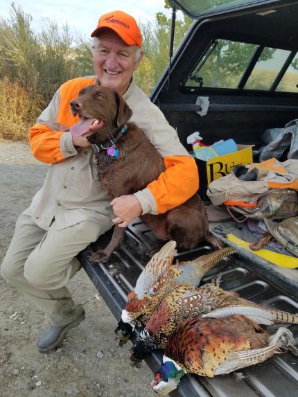 Photo of a hunter sitting on the tailgate of his care hugging his dog with three freshly harvested pheasants