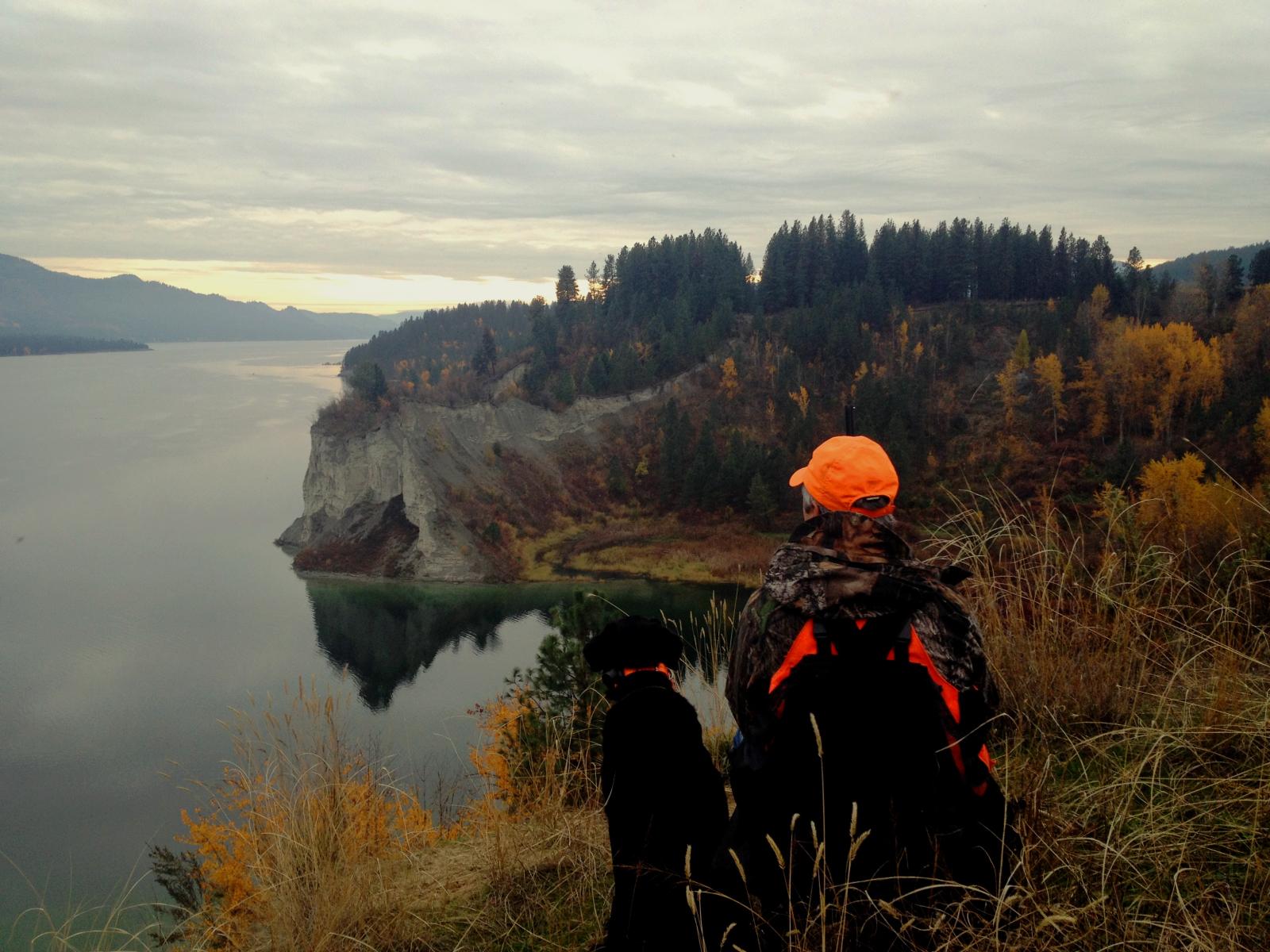 A hunter and their dog sitting on a high overlook in a wildlife area, with a lake and colorful fall trees in the distance