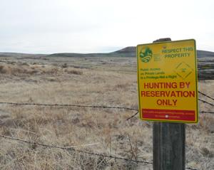 A yellow sign that reads "HUNTING BY RESERVATION ONLY" on a wood post in front of open range