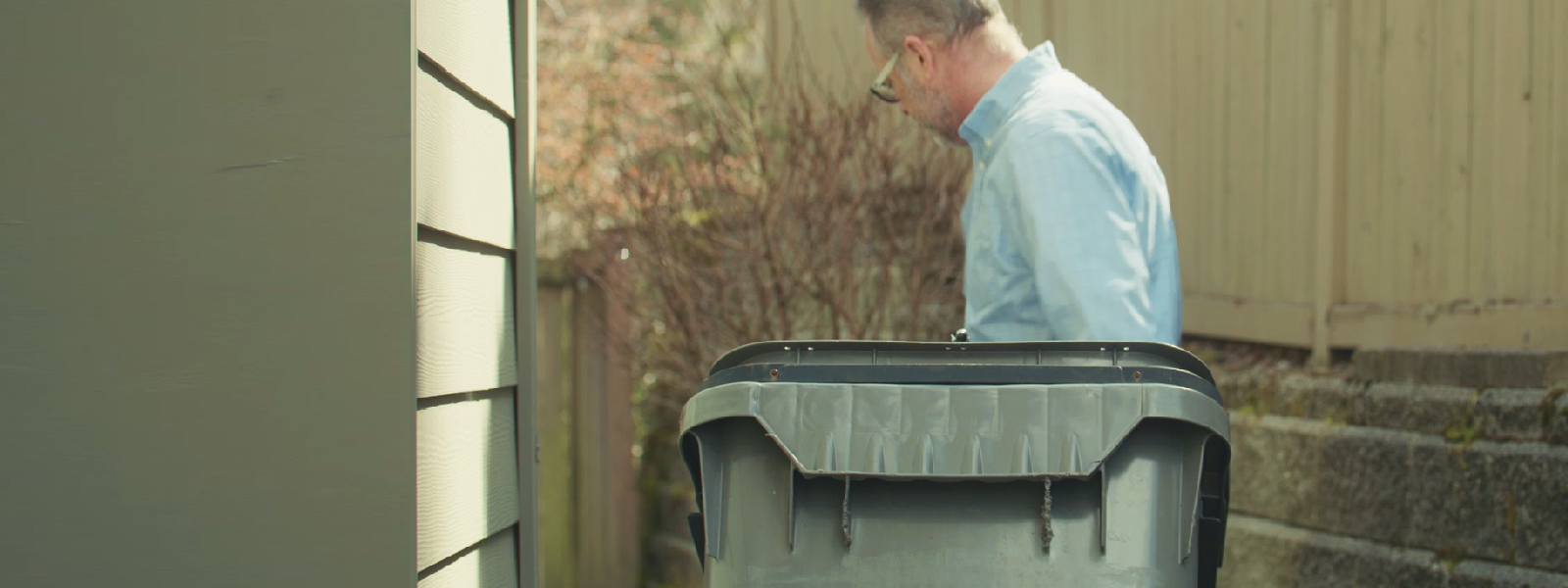 A man turned away from the camera, wheeling his garbage can next to his house
