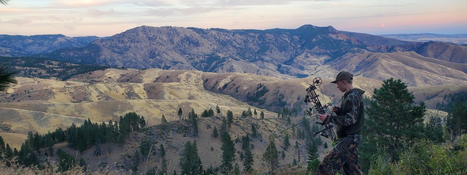 A young archery hunter with their bow standing on a ridge with an expansive canyon in the background