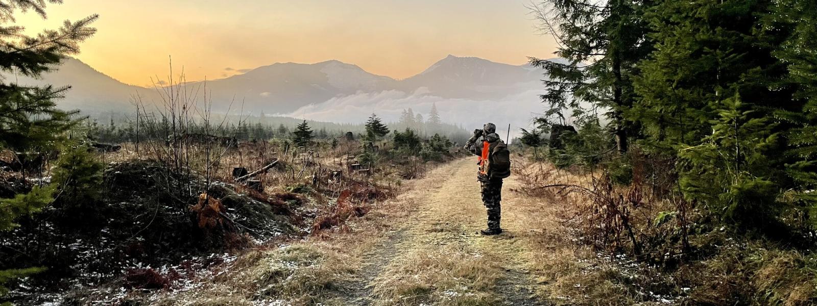 A hunter standing on a gravel road in the forest holding binoculars on a frosty morning
