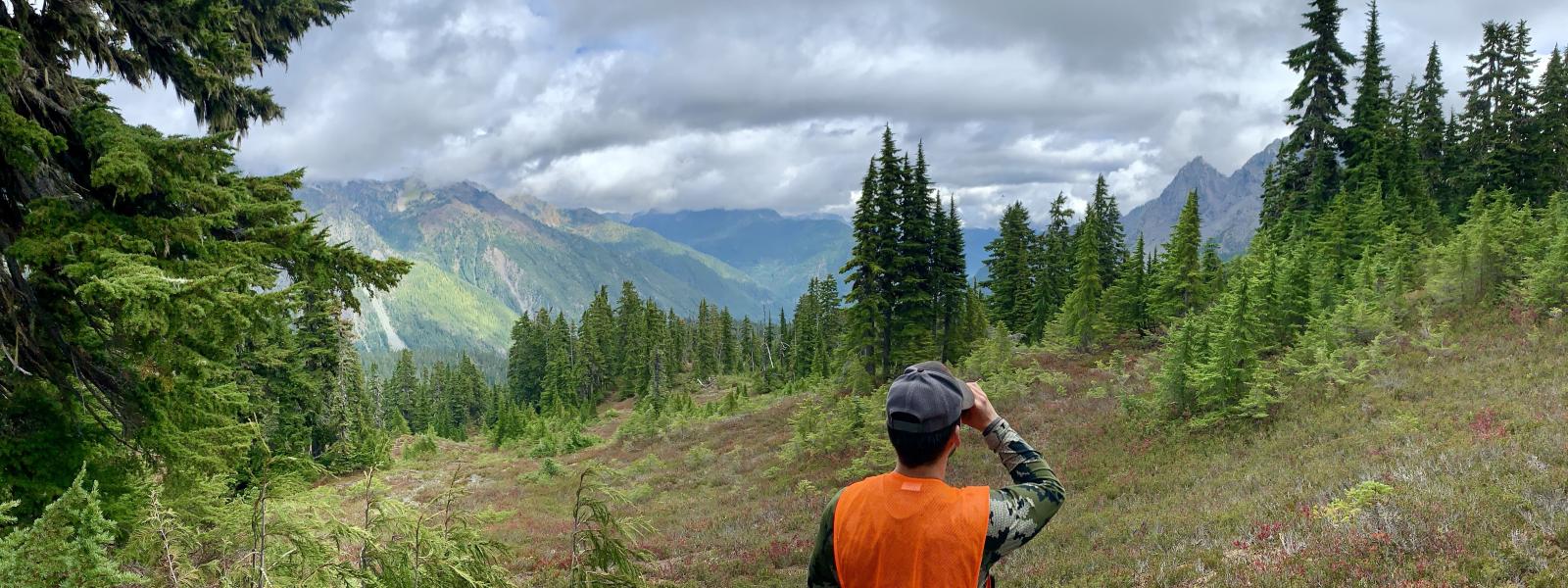 A hunter in an orange vest and a ball cap looking away from the camera towards a vast wilderness area