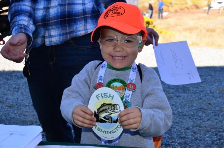 A child in a blaze orange hat holding up a WDFW sticker and smiling for the camera