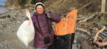 Woman wearing hat and binoculars holds up two bags of trash on beach.