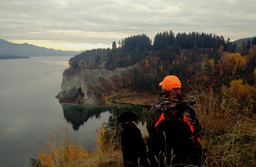 A hunter and their dog sitting on a high overlook in a wildlife area, with a lake and colorful fall trees in the distance