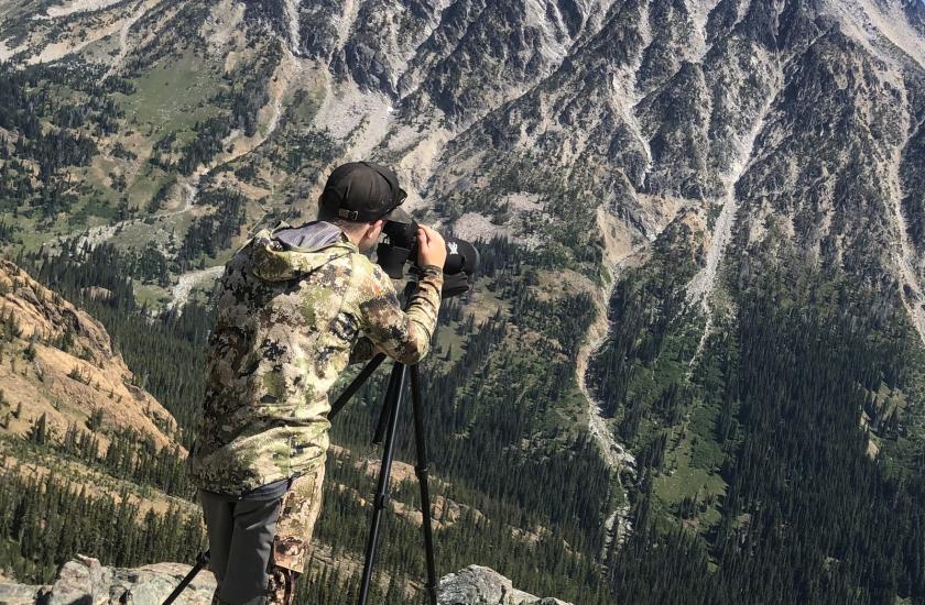 A hunter using a scope to scout for elk in a distant forest at the base of a mountain cliff