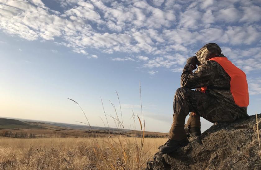 A hunter sitting on a rock in a field, using binoculars to look in the distance