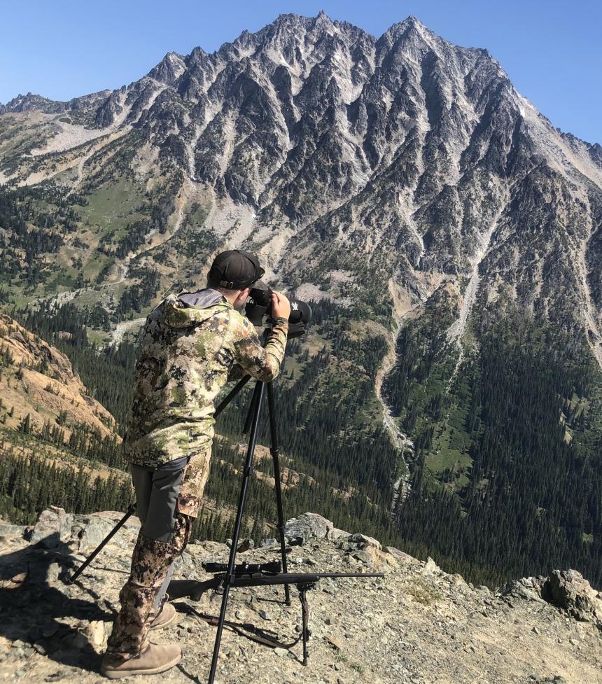 A hunter using a scope to scout for elk in a distant forest at the base of a mountain cliff