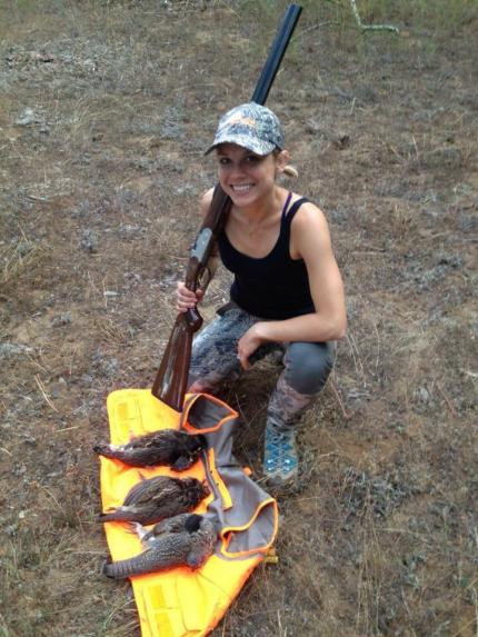 Young female hunter holding a shotgun and kneeling with three freshly harvested ruffed and blue grouse laying on an orange vest in the field.
