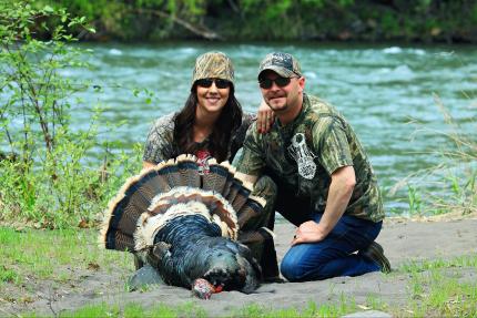 Photo of a man and women wearing camouflage knelling by a freshly harvested turkey with a river in the background. 