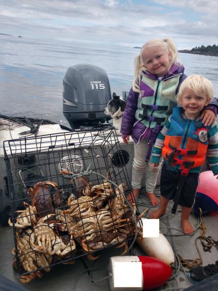 Two young children with life jackets stand next to full crab pot on a boat.