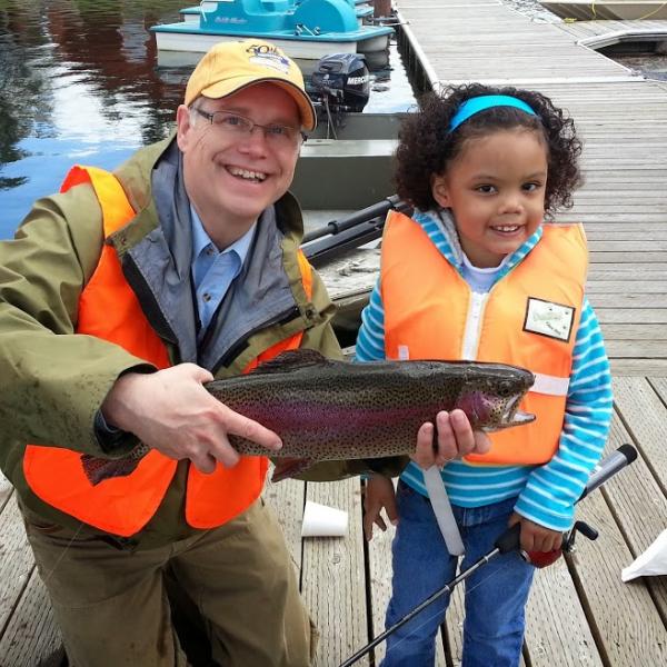 Volunteer holds a fish that was caught at youth fishing event, with young child standing nearby. 